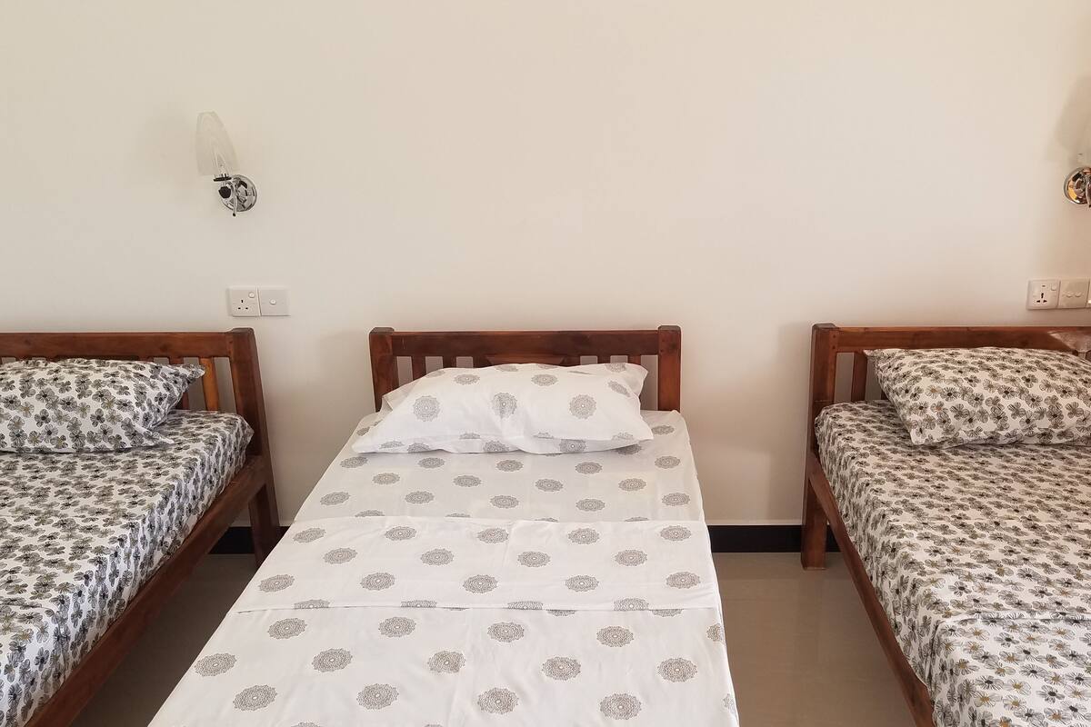 A room with two single beds, each with a floral-patterned bedspread and matching pillows. A central bed has a white bedspread with a geometric pattern. The room has wooden headboards and a plain white wall with two wall-mounted light fixtures.