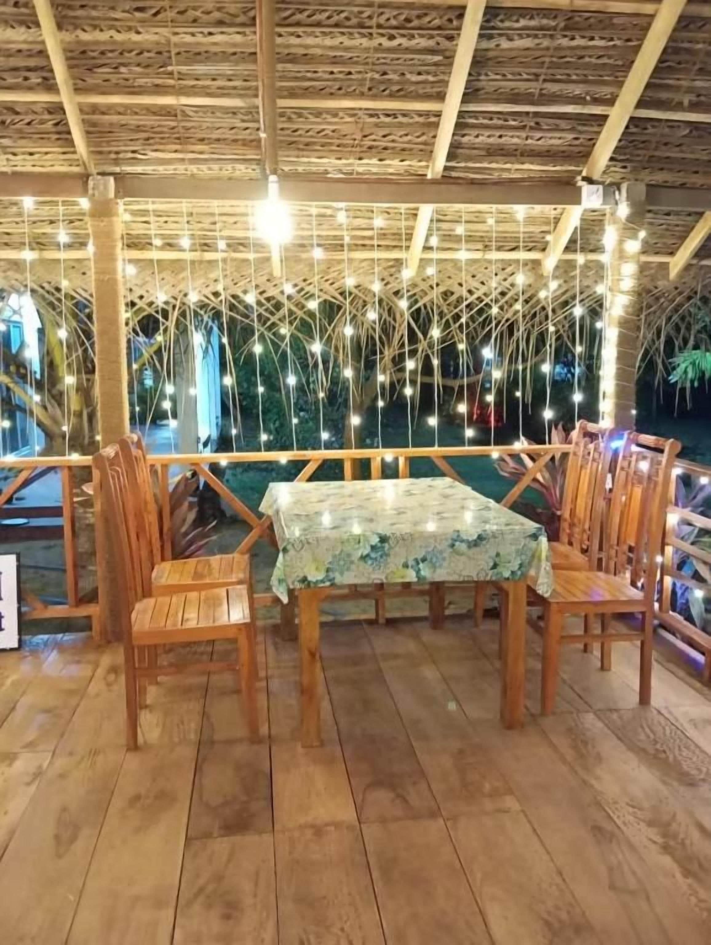 An outdoor dining area with wooden chairs and a table, under a thatched roof with string lights.