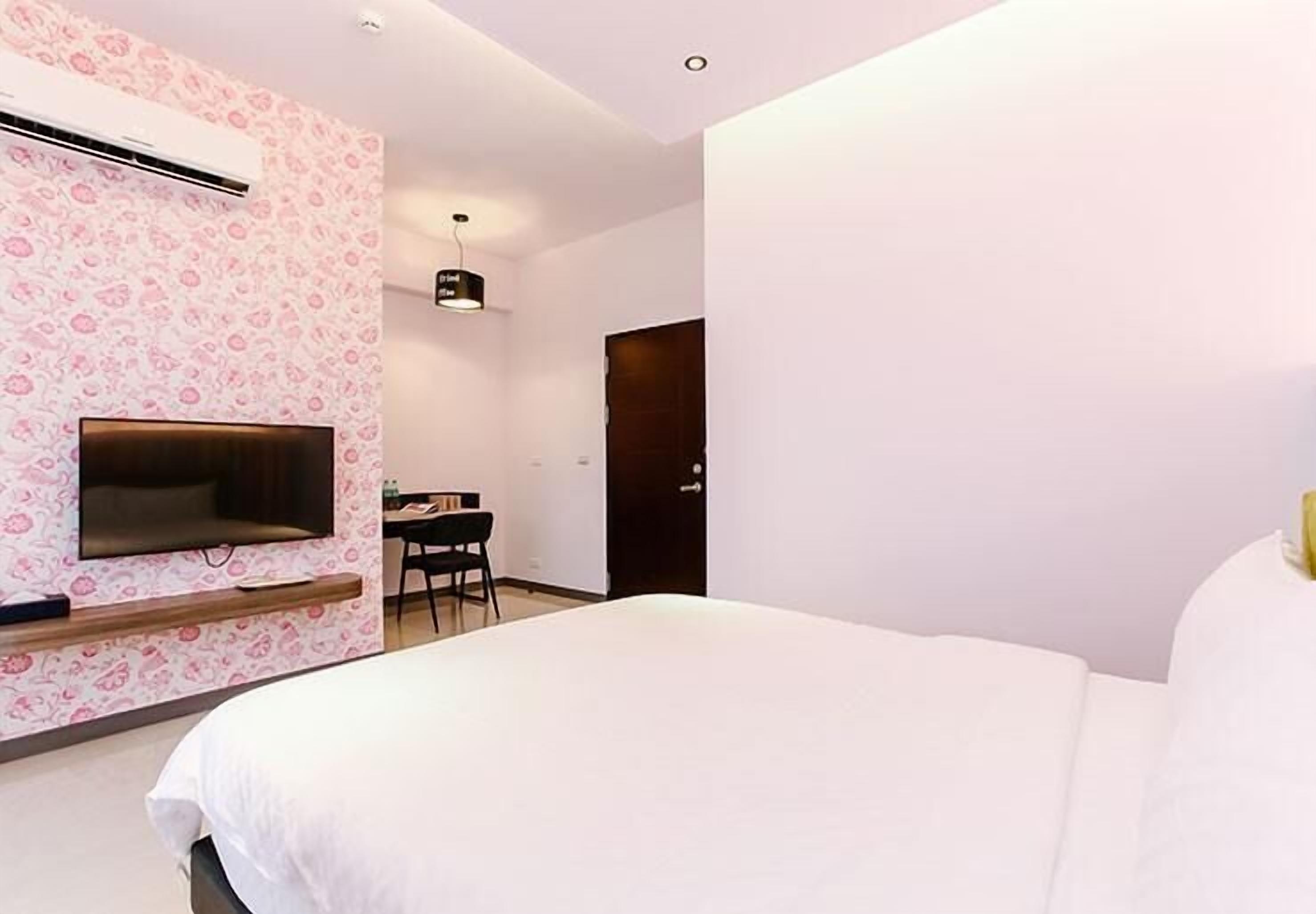 Ein modernes Hotelzimmer mit einem großen Bett, einem Flachbildfernseher an einer Wand mit floralem Muster, einem Schreibtisch mit Stuhl und einer Tür im Hintergrund.