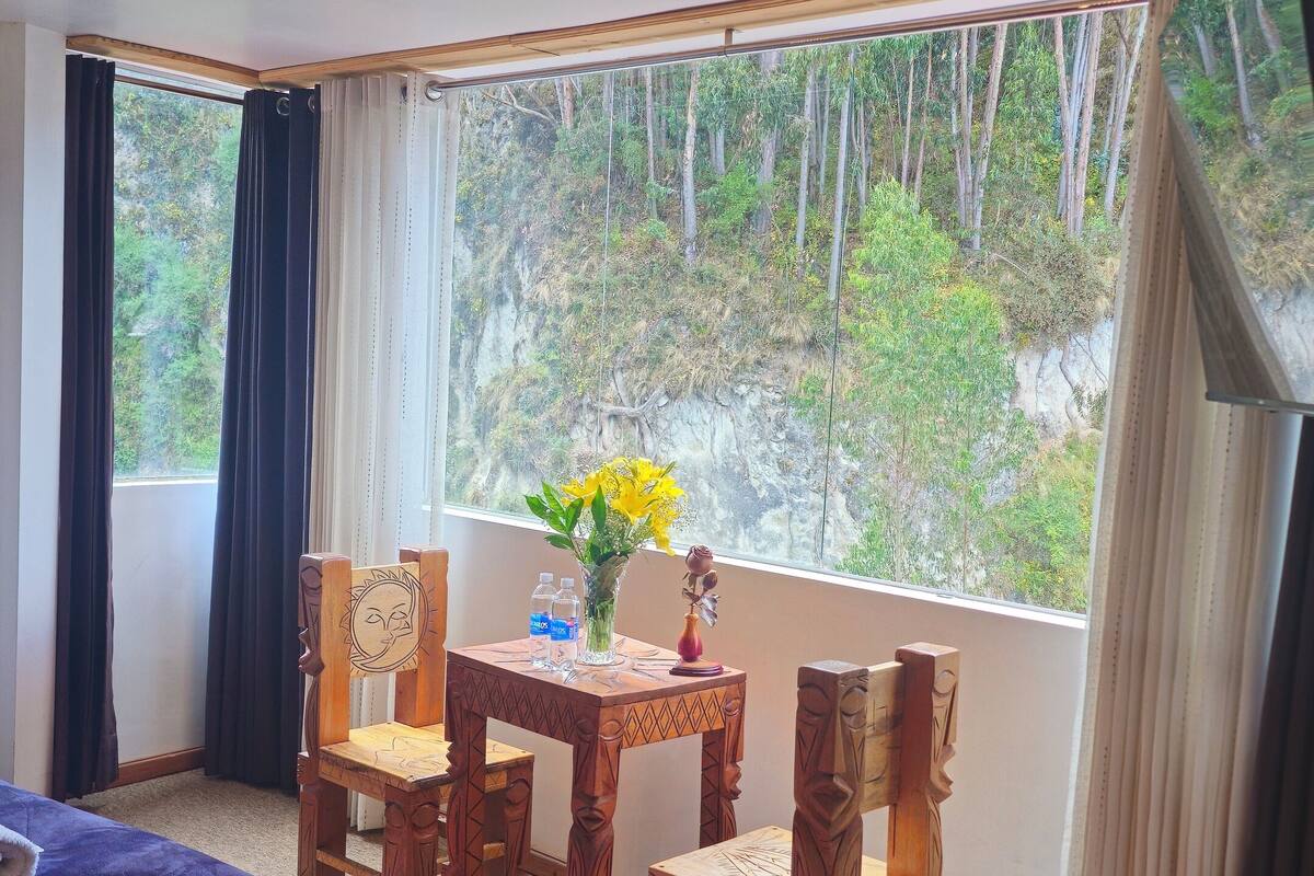 Ein Zimmer mit zwei Betten, einem Holztisch mit Stühlen, einer Vase mit Blumen und Blick auf einen Wald durch ein großes Fenster.