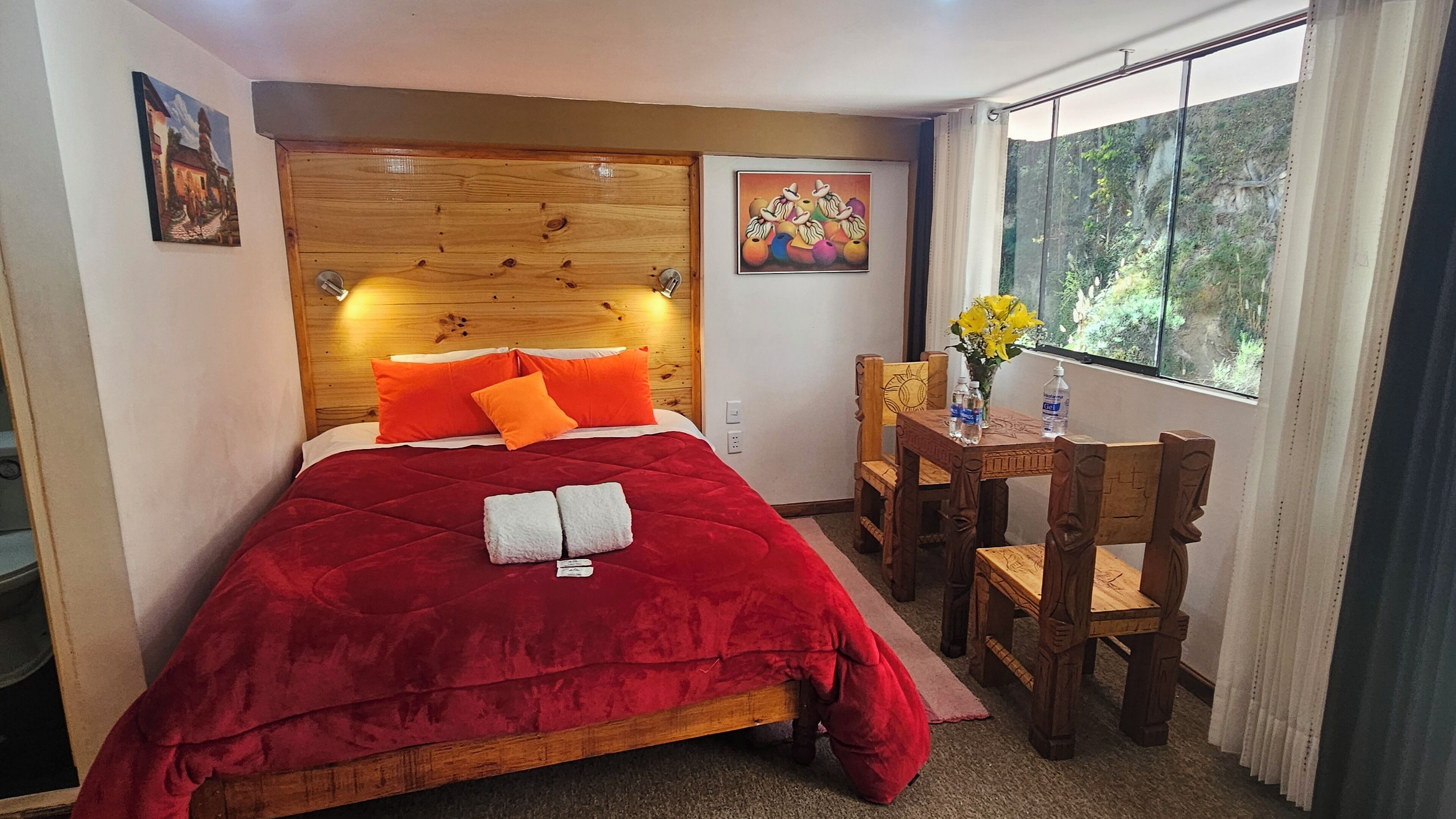 A bedroom with a wooden headboard, a red bedspread, and a wooden chair with a table beside the window.