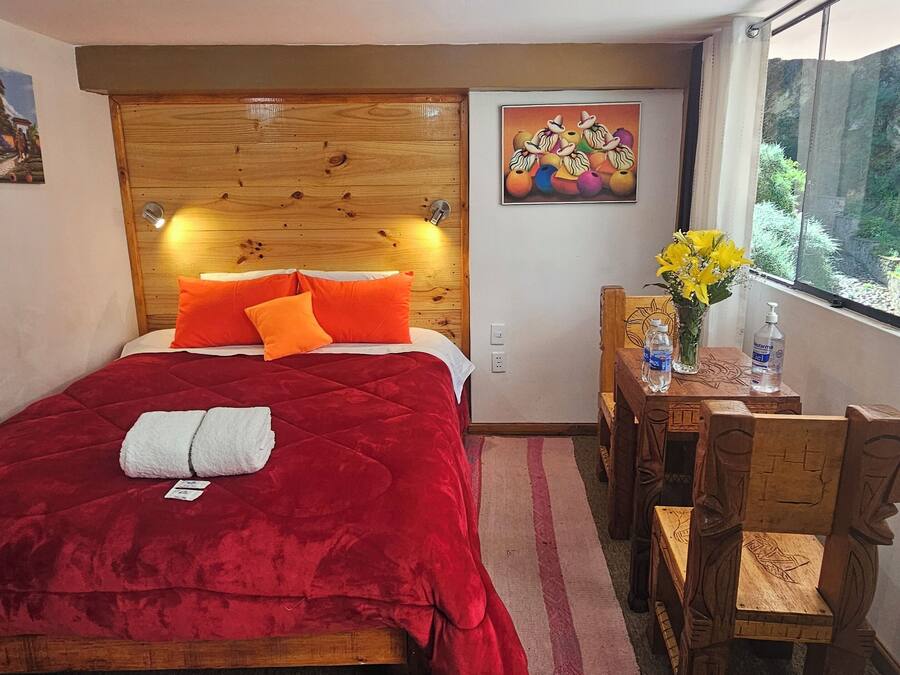 Un dormitorio con cabecera de madera, colcha roja, dos almohadas naranjas, toalla blanca, mesita de noche con un jarrón de flores y ventana con vistas a la vegetación.