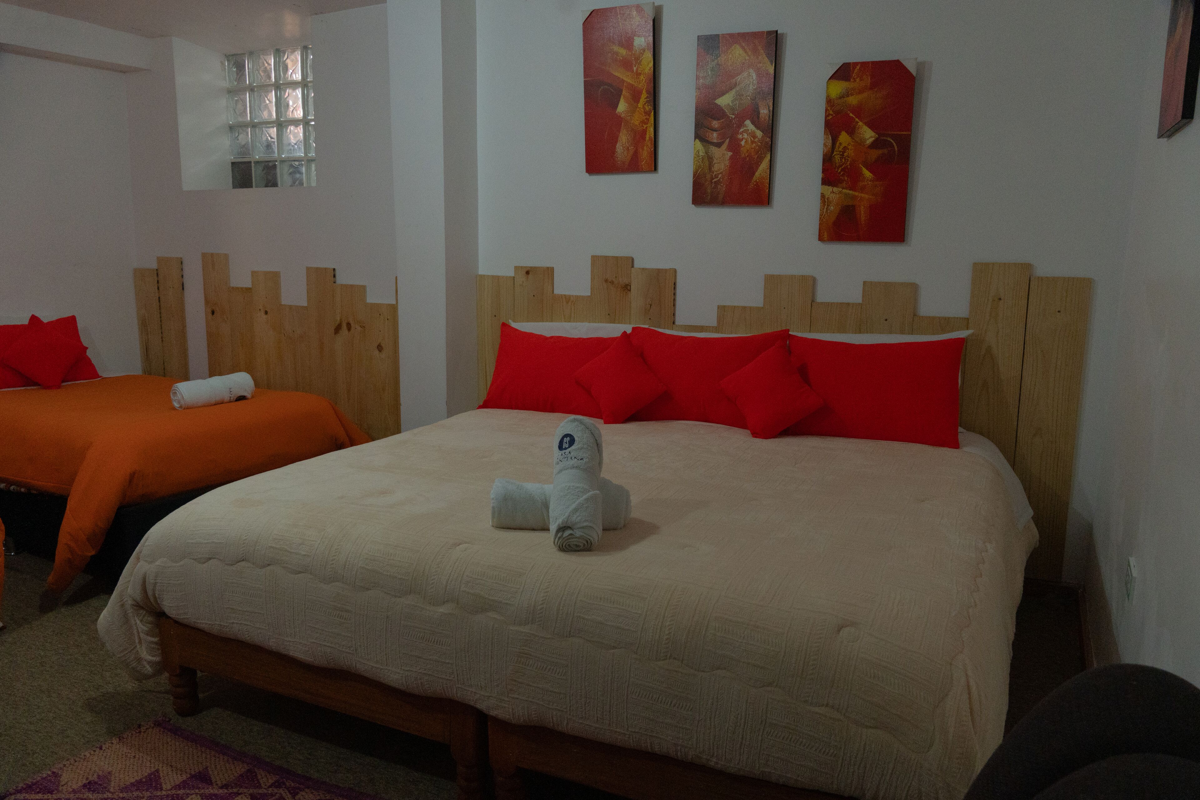 A hotel room with two beds, one with a beige bedspread and the other with an orange bedspread, both with wooden headboards and red pillows.