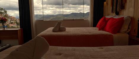 Habitación de hotel con un ventanal grande que ofrece vistas a la montaña, una cama con ropa de cama roja y blanca, y un cabecero de madera.