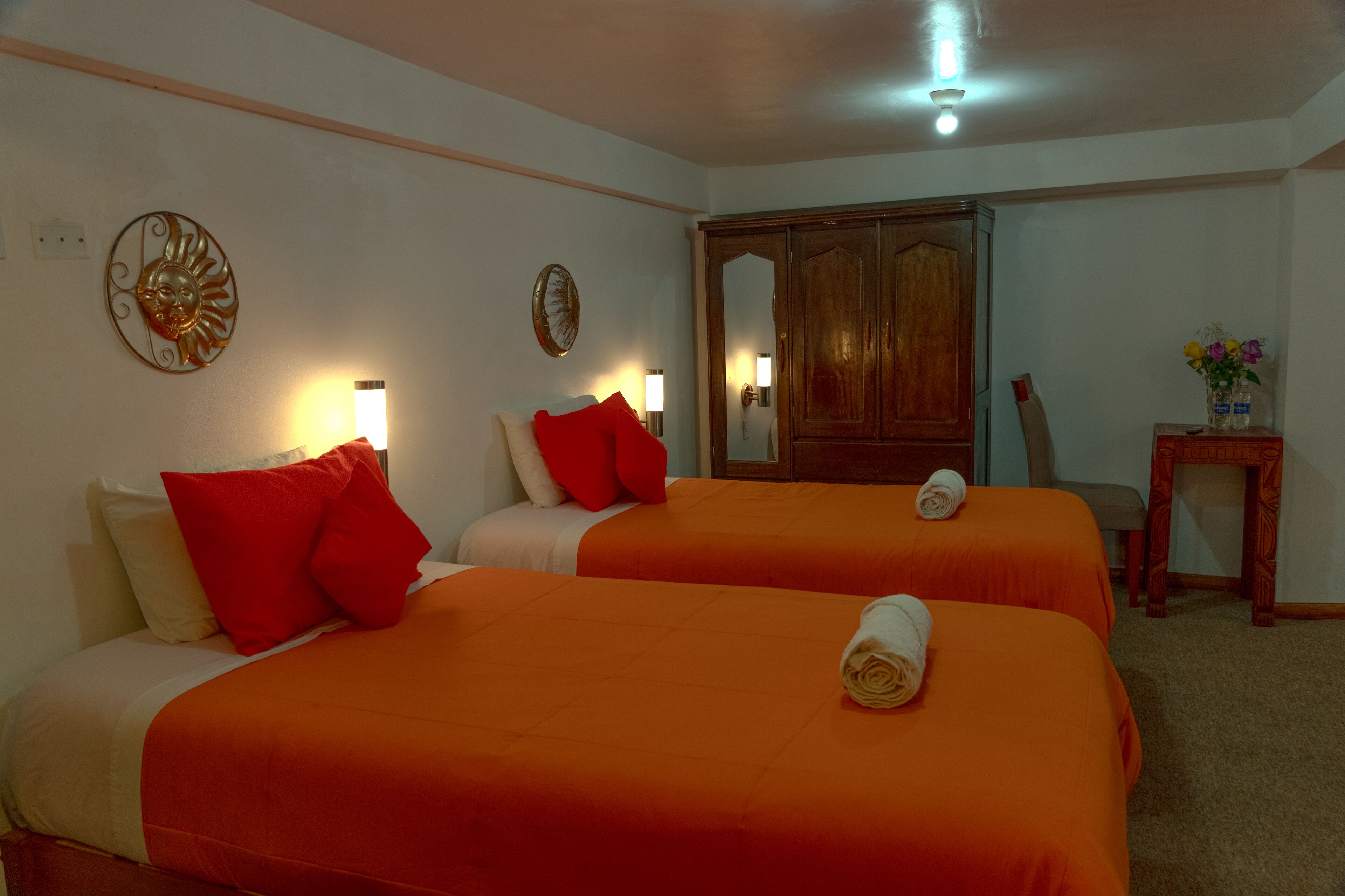 Ein Hotelzimmer mit zwei Betten, jeweils mit orangefarbener Bettwäsche und roten Kissen. Es gibt Wandleuchten, einen Holzschrank und einen kleinen Tisch mit einem Blumenstrauß.