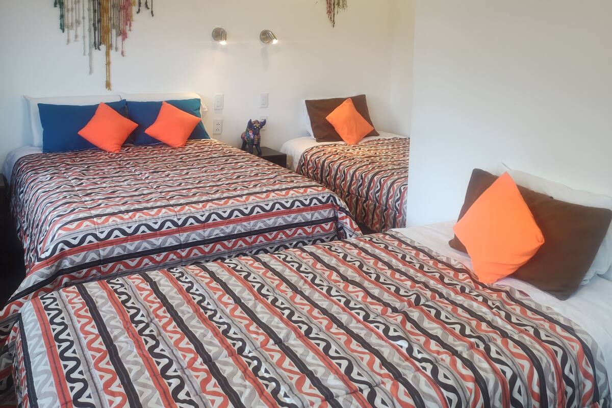 Ein Zimmer mit zwei Betten, jeweils mit gemusterten Bettbezügen und orangefarbenen Kissen. An den Wänden sind Leuchten angebracht, und über den Betten hängt ein dekoratives Element.
