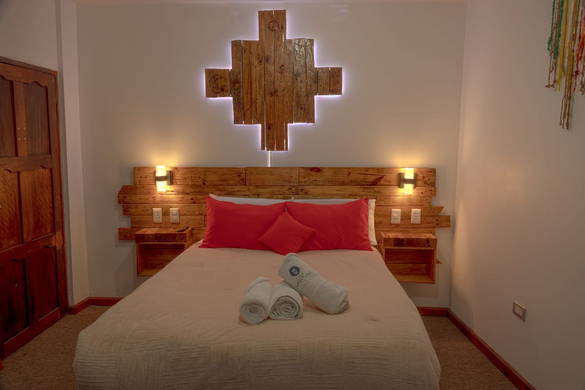Ein Hotelzimmer mit einem hölzernen Kopfteil, einer kreuzförmigen Leuchte, zwei Nachttischen, einem Bett mit roten Kissen und zwei aufgerollten Handtüchern.