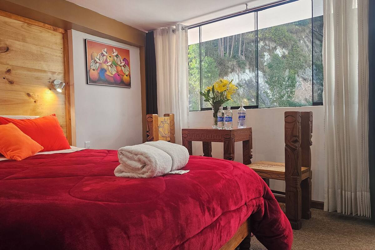Ein Bett mit roter Bettwäsche, ein Holz-Kopfteil, ein Bild an der Wand, ein Fenster mit Blick auf Bäume und ein Tisch mit Wasserflaschen.