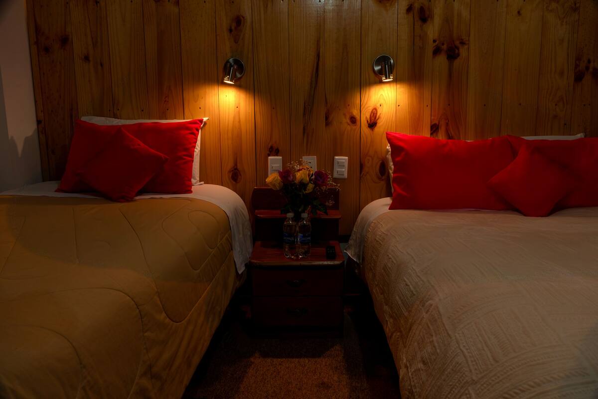 Ein Zimmer mit zwei Betten, jedes mit einem roten Kopfkissen, Holzwänden und Nachttischlampen.