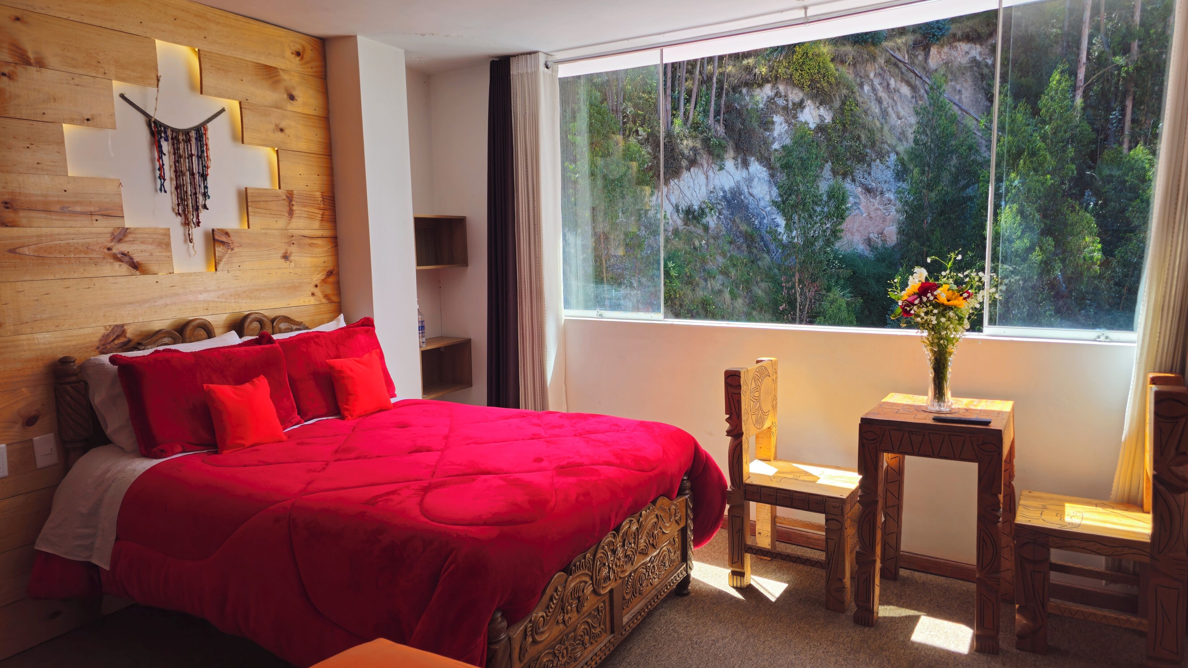 Un dormitorio con una cama grande, un cabecero de madera, una mesita de noche, una silla, una pequeña mesa y una ventana con vistas a un bosque.