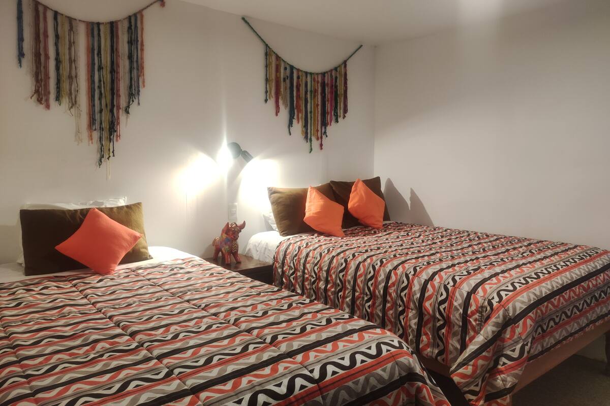 Ein Schlafzimmer mit zwei Betten, jeweils mit gemusterten Bettbezügen und orangefarbenen Kissen. An den Wänden sind Lampen angebracht und auf dem Nachttisch steht ein kleines Dekorationsstück.