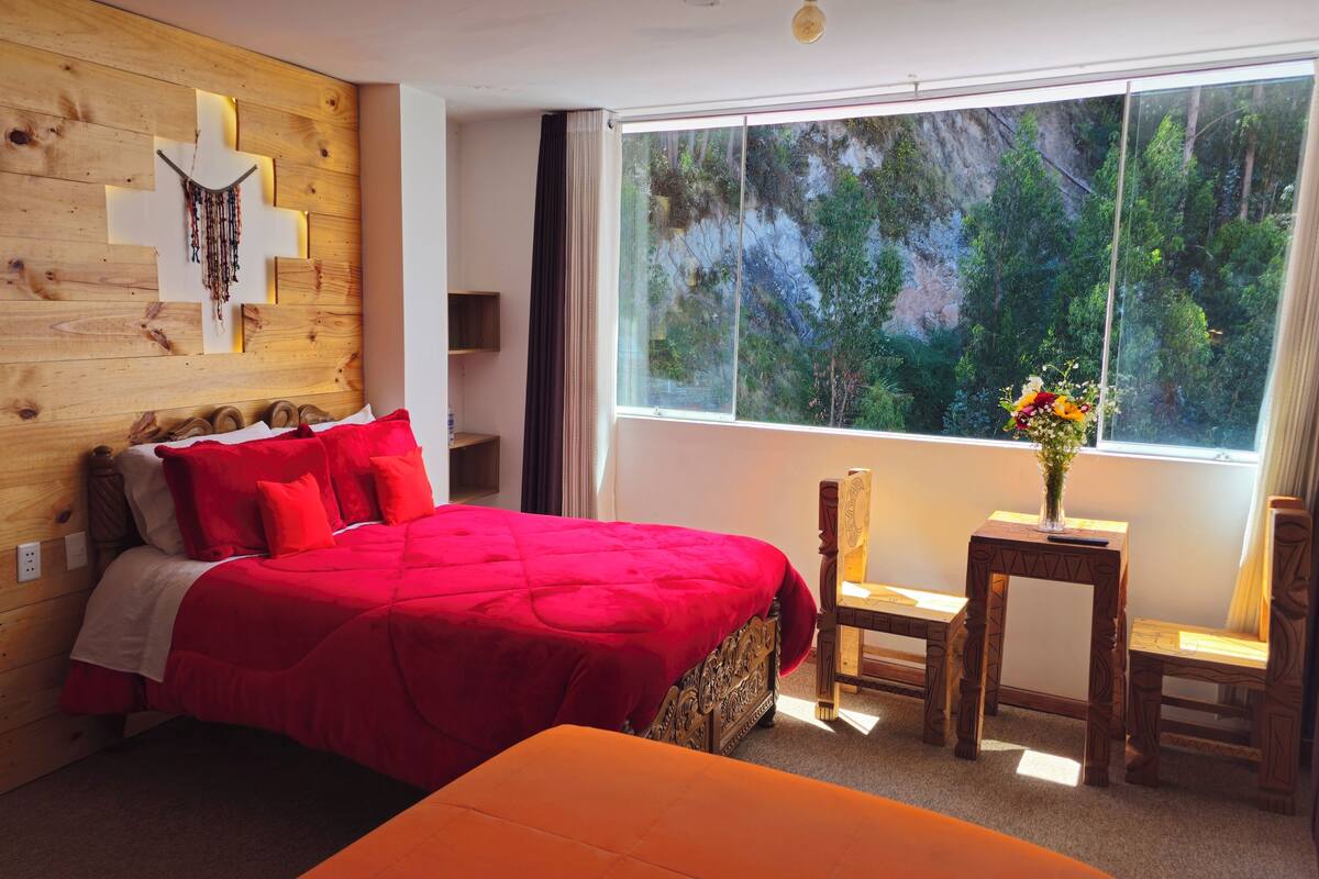 Ein Schlafzimmer mit einem hölzernen Kopfstück, einer roten Bettdecke, einem kleinen Tisch mit einer Vase und Blick ins Grüne durch ein Fenster.