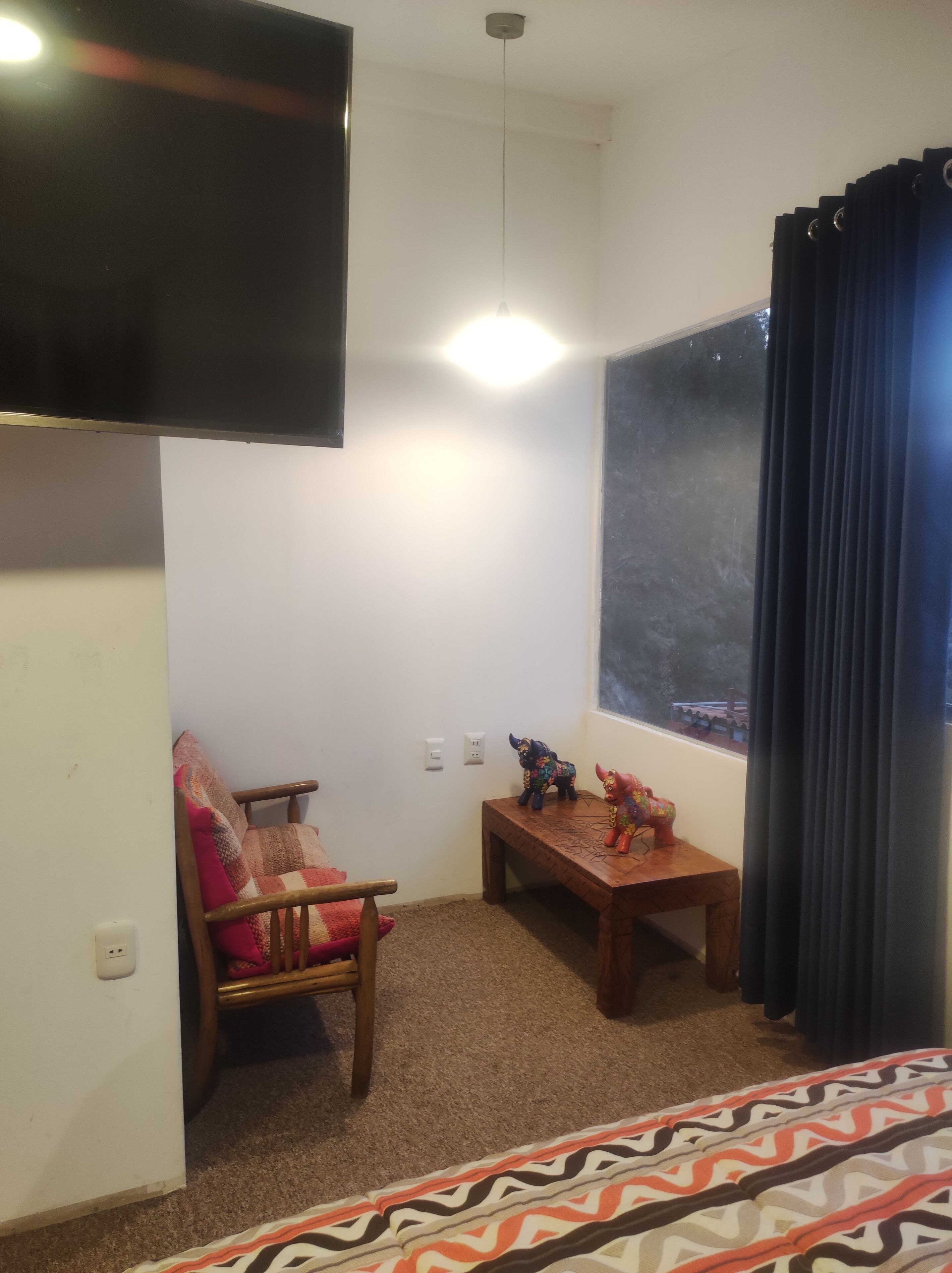 A room with a flat-screen TV mounted on the wall, a wooden bench, a chair with a colorful cushion, and a window with curtains.