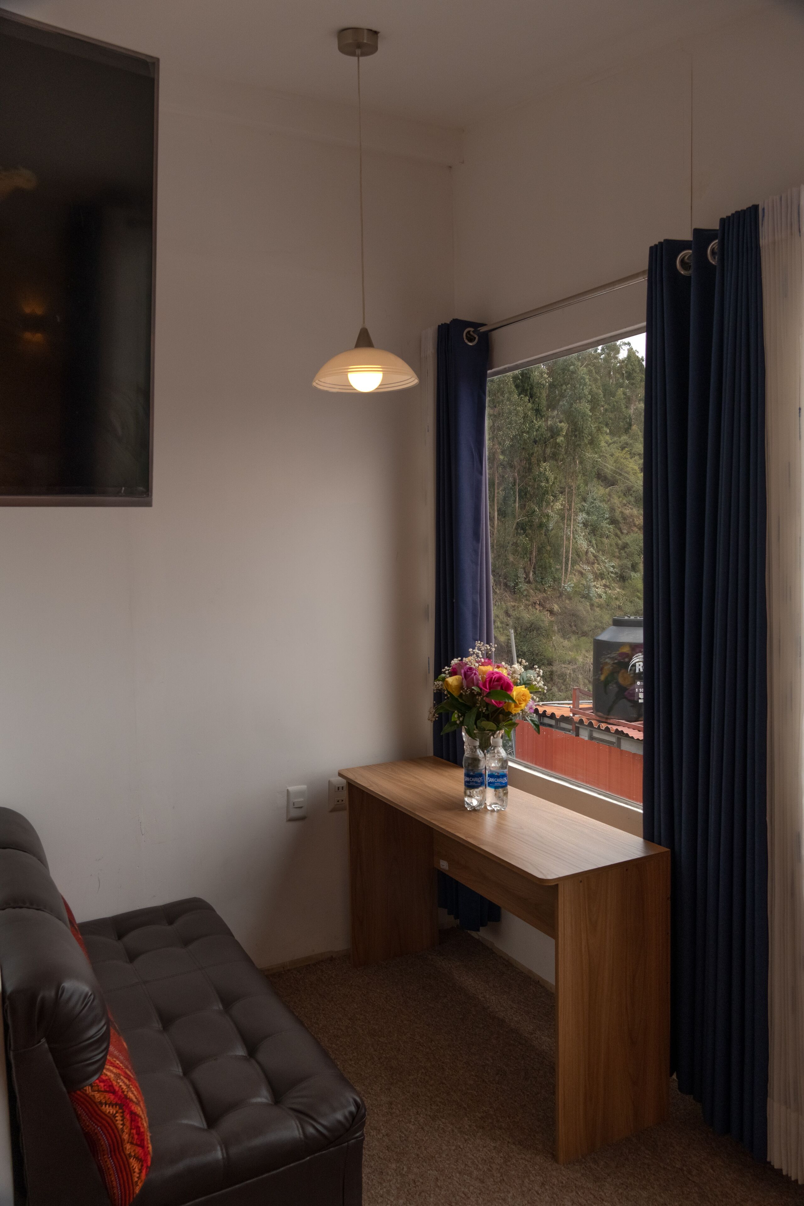 Una habitación con un escritorio de madera, un sofá de cuero negro, una lámpara colgante y una ventana con cortinas.
