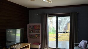 Een woonkamer met een flatscreen televisie, houten vloeren, een plafondventilator, een boekenkast en een glazen schuifdeur die leidt naar een buitenruimte.