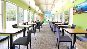 Ein Restaurant mit langen Tischen und Stühlen, großen Fenstern und einem Wandgemälde.