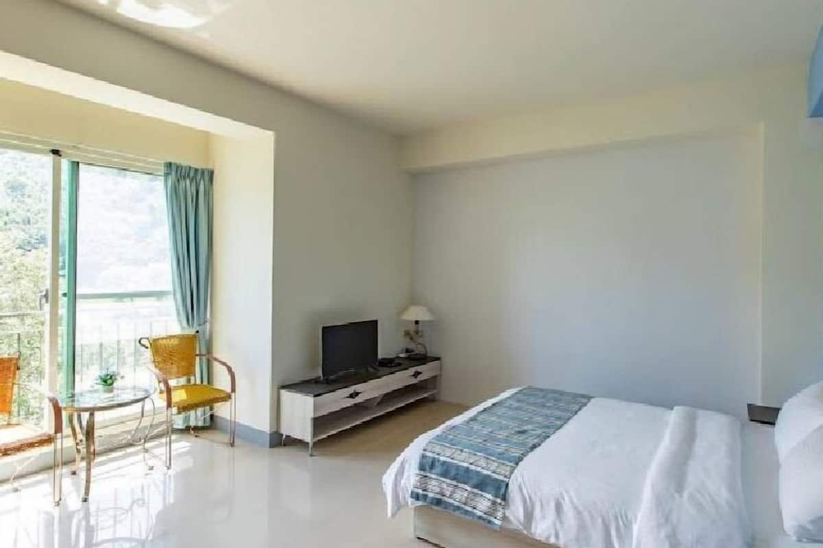 Quarto com cama, televisão em móvel, cadeira e janela com cortinas.
