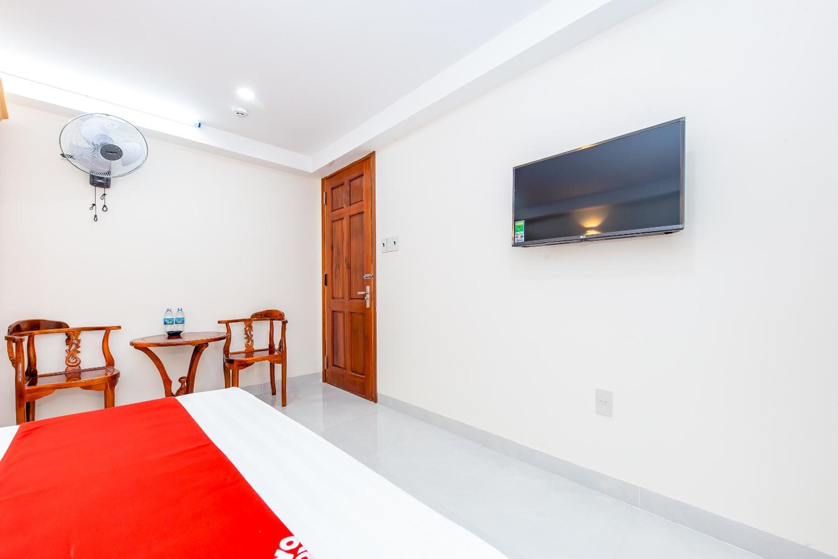 Una habitación de hotel con una cama, un televisor montado en la pared, un ventilador de techo y dos sillas de madera.