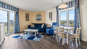 Una sala de estar con un sofá azul, una mesa de comedor con sillas, una mesa de centro y vistas de la ciudad a través de grandes ventanales.