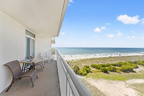 A balcony with wicker chairs and a table, overlooking a beach with people and umbrellas.