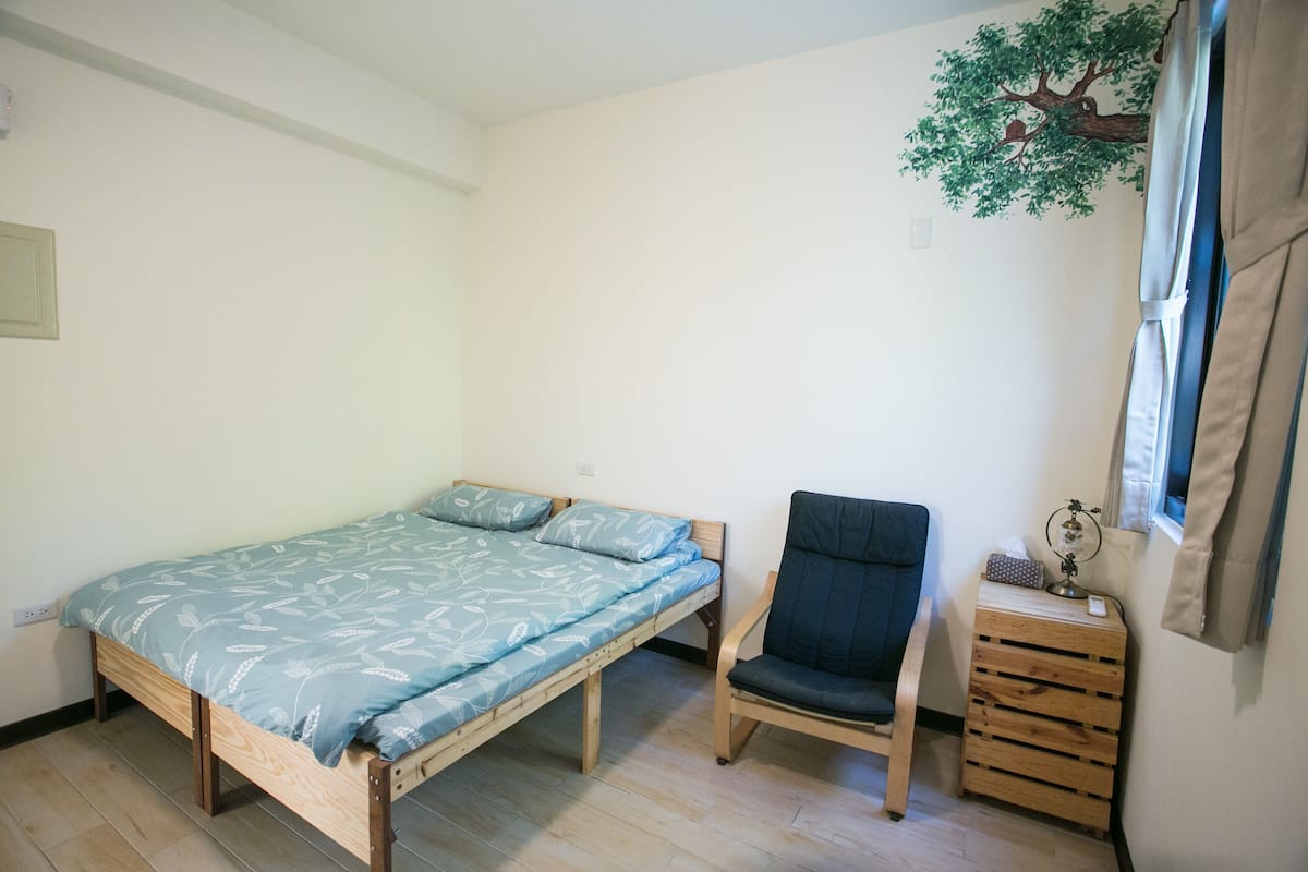 A small, single-bed room with a wooden bed frame, a dark chair, and a small wooden side table.
