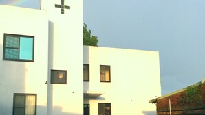 A modern white building with a cross on the facade, a small porch, and a garden area with plants and a red chair. - Tree House Café & Homestay (Tongluo)
