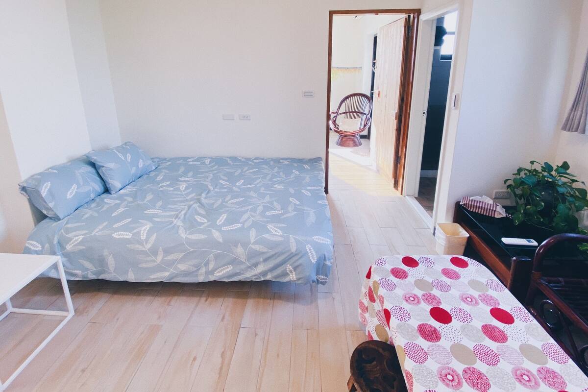 A small, bright room with a bed, a desk, and a chair. There is a door leading to another room.