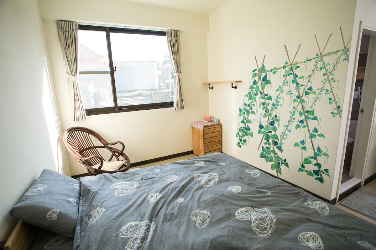 A bedroom with a bed, a chair, a window with curtains, a dresser, and a wall-mounted plant.