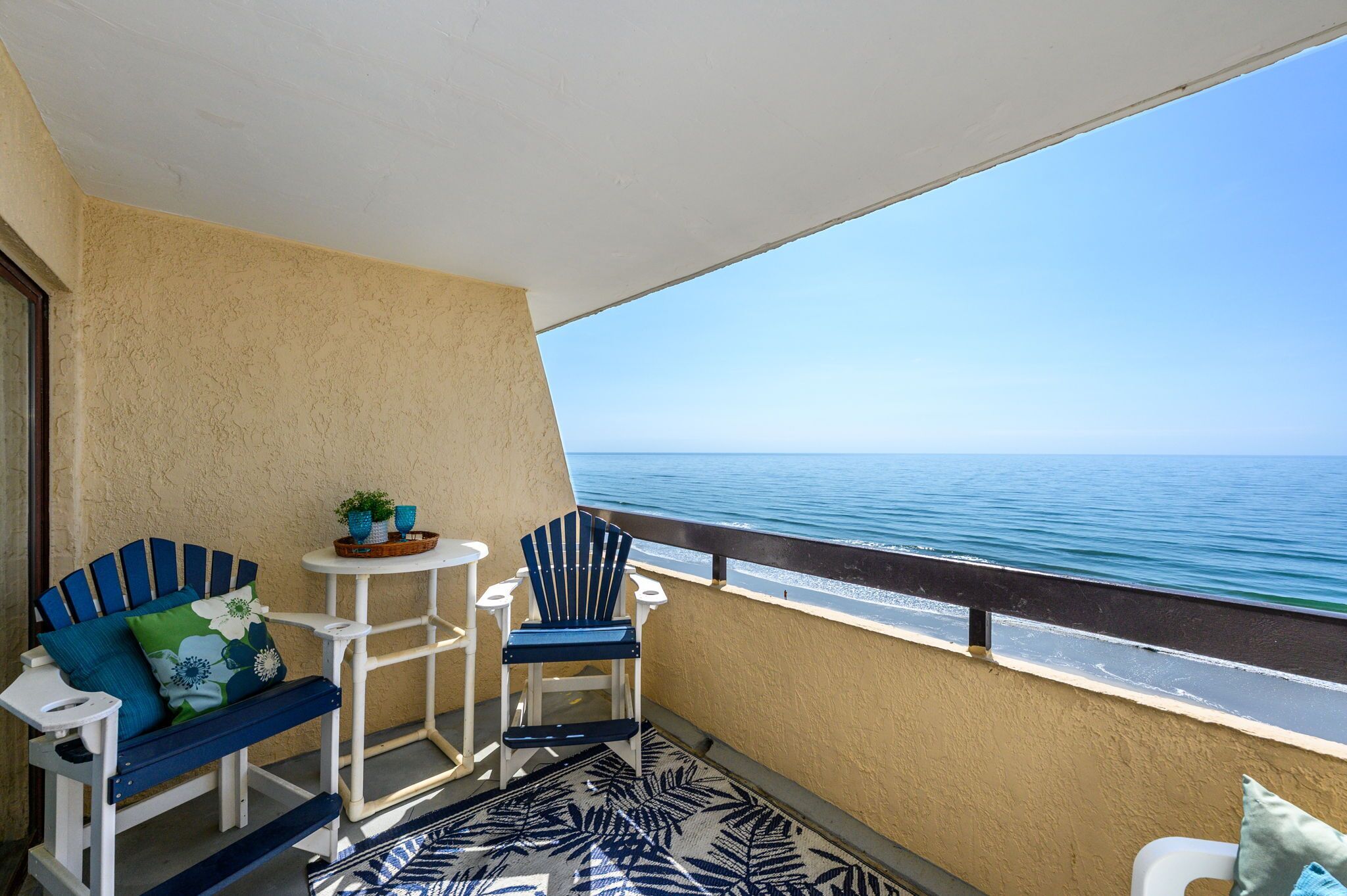 A balcony with a view of the ocean, a blue and white patterned rug, a white table with a basket, two blue chairs, and a blue sofa with pillows.
