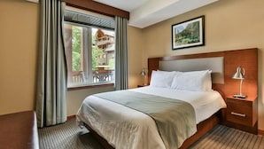 A hotel room with a large bed, a wooden headboard, a nightstand with a lamp, and a window with curtains.