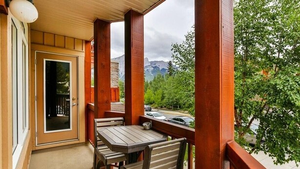 A wooden balcony with a table and chairs, a door, and a view of a road and greenery.