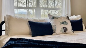 Una cama con ropa de cama blanca y azul oscuro, una manta azul oscuro, y una ventana con vistas a árboles y montañas.