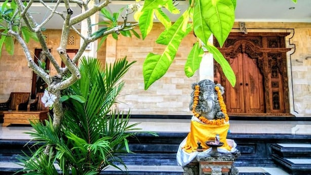 A statue of a deity with a yellow cloth, surrounded by greenery and a stone base, in front of a wooden door with intricate carvings. - Santi HomeStay (Canggu)