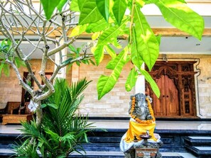 Eine Statue einer Gottheit mit gelbem Stoff, umgeben von Grün und einem steinernen Sockel, vor einer Holztür mit aufwendigen Schnitzereien.