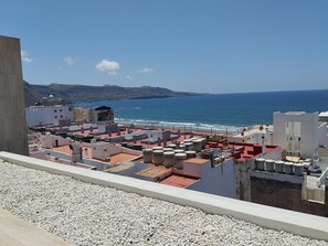 A rooftop terrace with a view of the sea and mountains, featuring buildings with red and white roofs and a gravel ground.