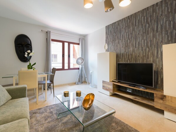A modern living room with a glass coffee table, a sofa, a TV on a wooden stand, and a patterned wall.