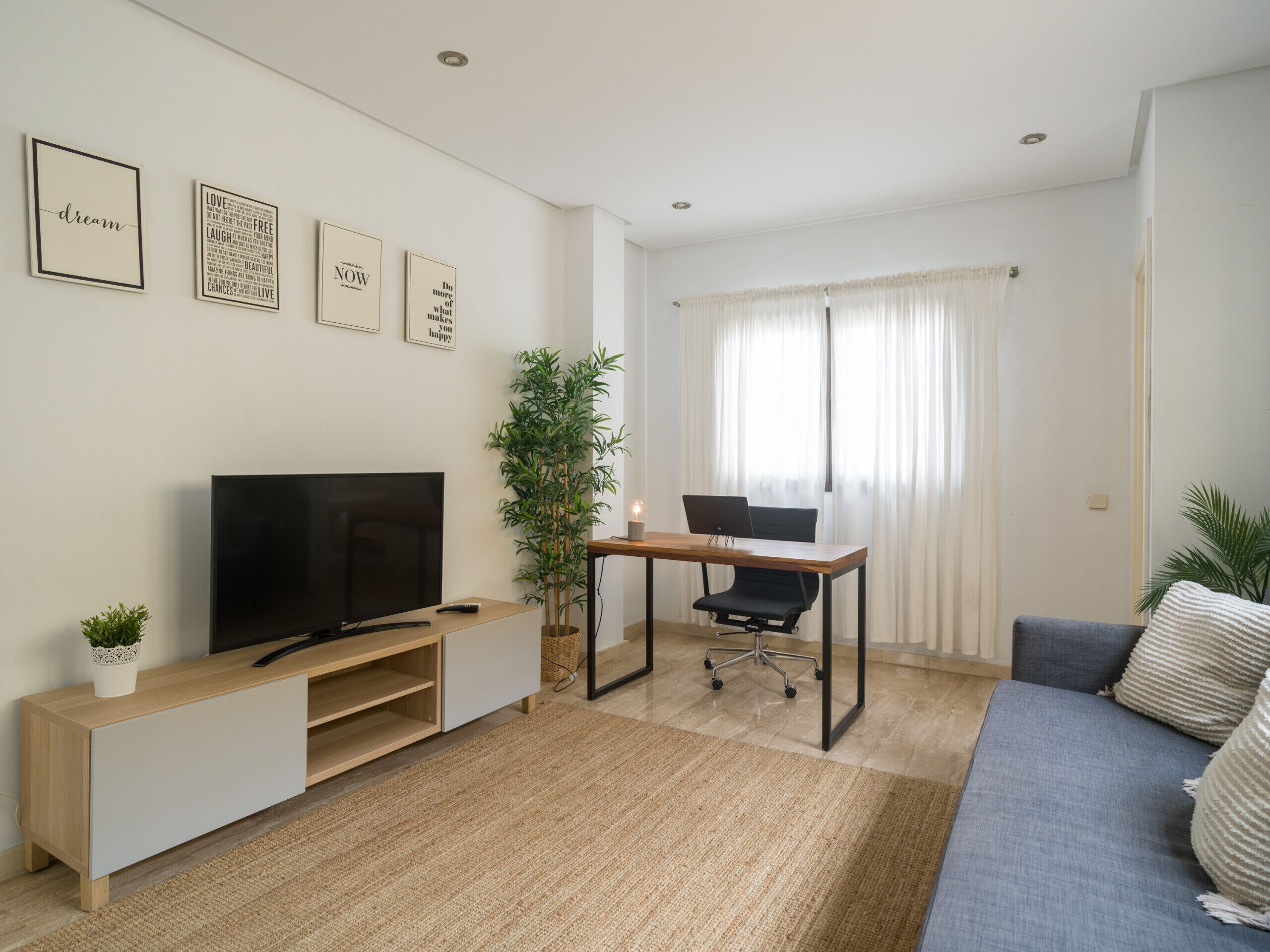 Una sala de estar moderna con una televisión, un escritorio con una computadora portátil, un sofá y un amplio ventanal con cortinas.