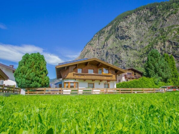 A chalet-style building with a sloped roof, wooden exterior, and a balcony. It is surrounded by lush green fields and a mountain backdrop. - Apartment Anna by Interhome (Umhausen)