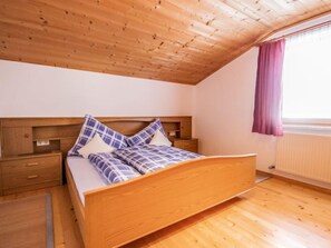 A bedroom with a wooden ceiling, a bed with plaid bedding, a nightstand, and a window with a curtain. - Apartment Anna by Interhome (Umhausen)