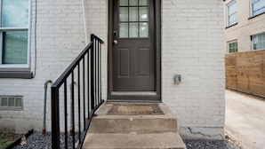 A front view of a house with a brown door, white brick walls, a black metal railing, and a concrete step leading to the door. - Beautiful, Renovated, Cozy & Quiet Vacation Home in Central Houston! (Houston)