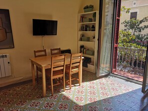 A dining area with a wooden table and chairs, a flat-screen TV mounted on the wall, a bookshelf with various items, and a patterned tile floor.