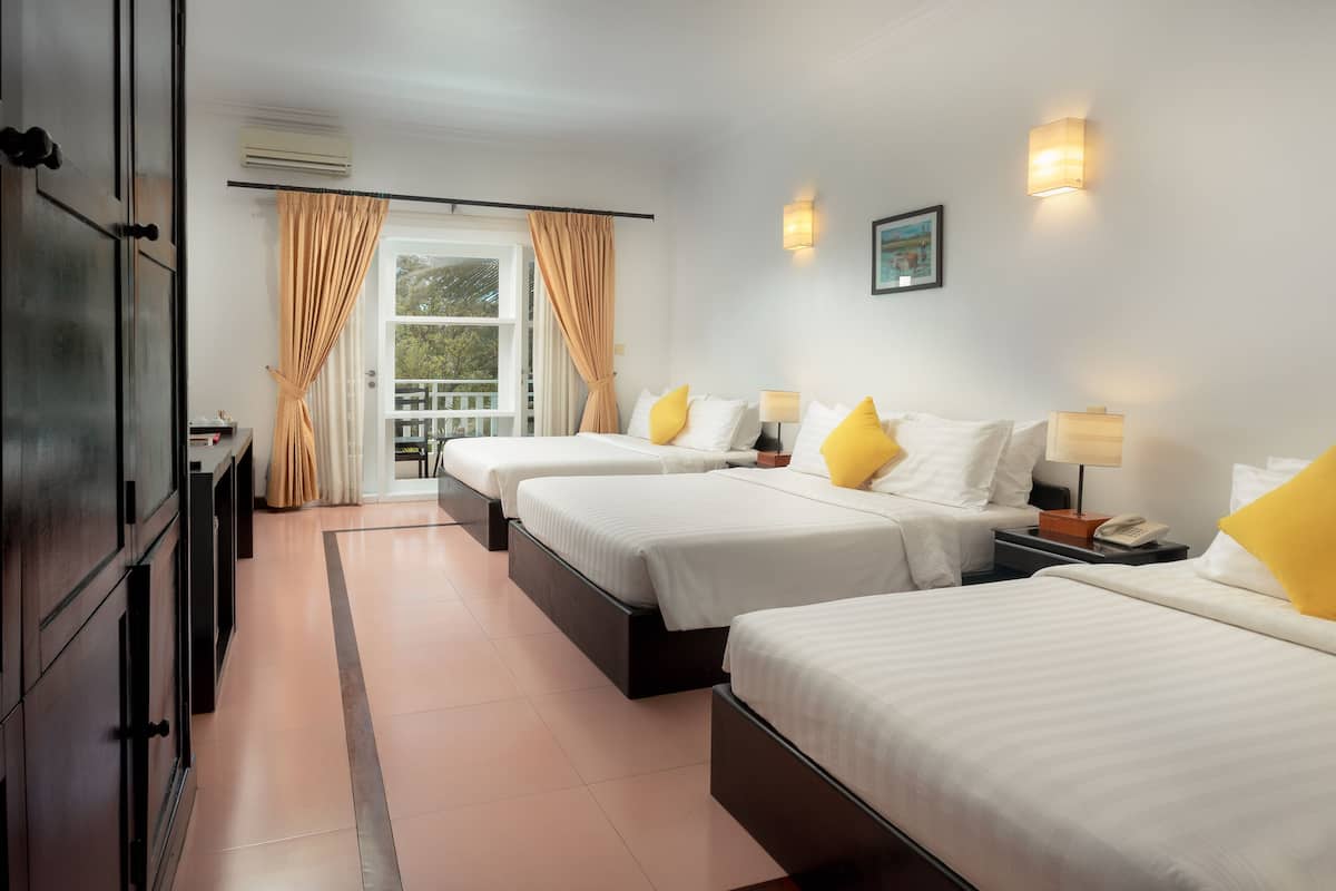 A hotel room with three beds, a large window with curtains, a wall-mounted air conditioner, and a small table with a lamp.