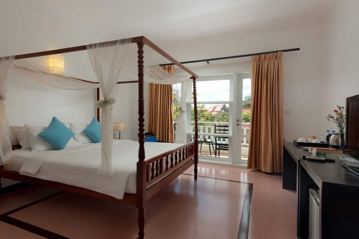 A four-poster bed with white linens and blue pillows, a bedside table with a lamp, and a view of the balcony.