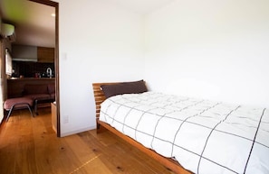 A bedroom with a wooden bed, a dark headboard, and a white quilt. A small kitchenette is visible in the background. - Mino (Mitoyo)