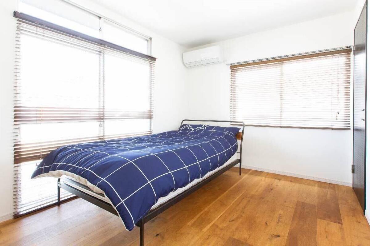 Una cama individual en una habitación con piso de madera, paredes blancas y dos ventanas con persianas.