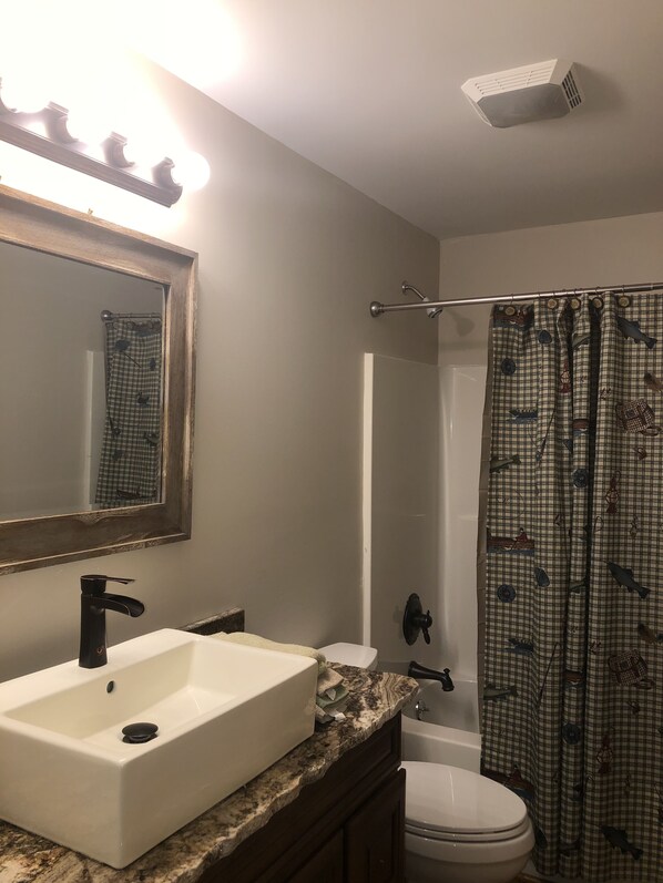 Un baño con lavamanos blanco, un espejo, una cortina de ducha con motivos náuticos y un ventilador en el techo.
