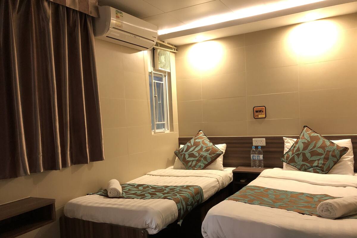 A hotel room with two beds, a window, an air conditioning unit, a bedside table, and a wall-mounted WiFi sign.