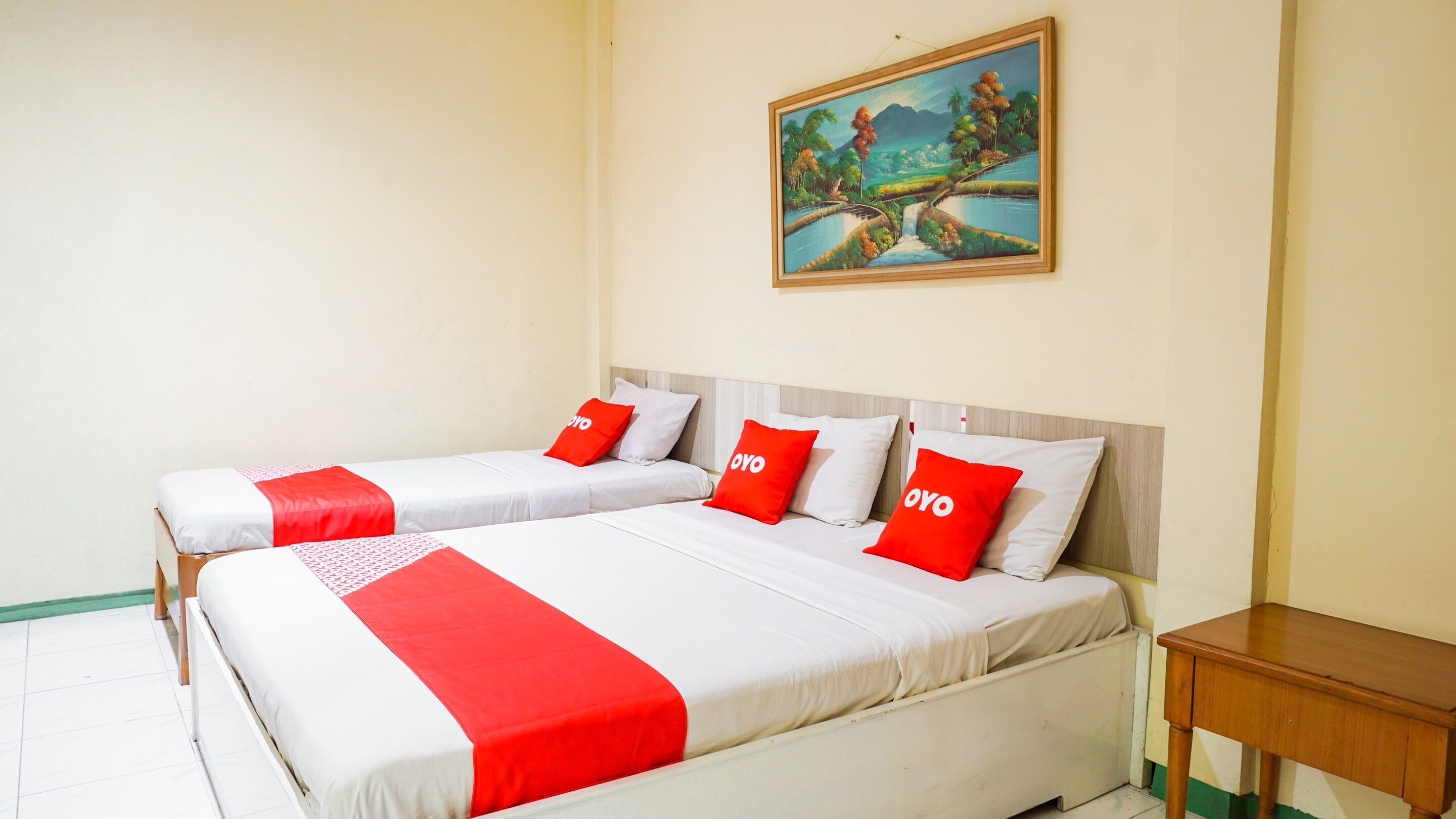 Habitación de hotel con dos camas, cada una con ropa blanca y detalles en rojo, un cuadro de un paisaje en la pared y una pequeña mesa de madera.
