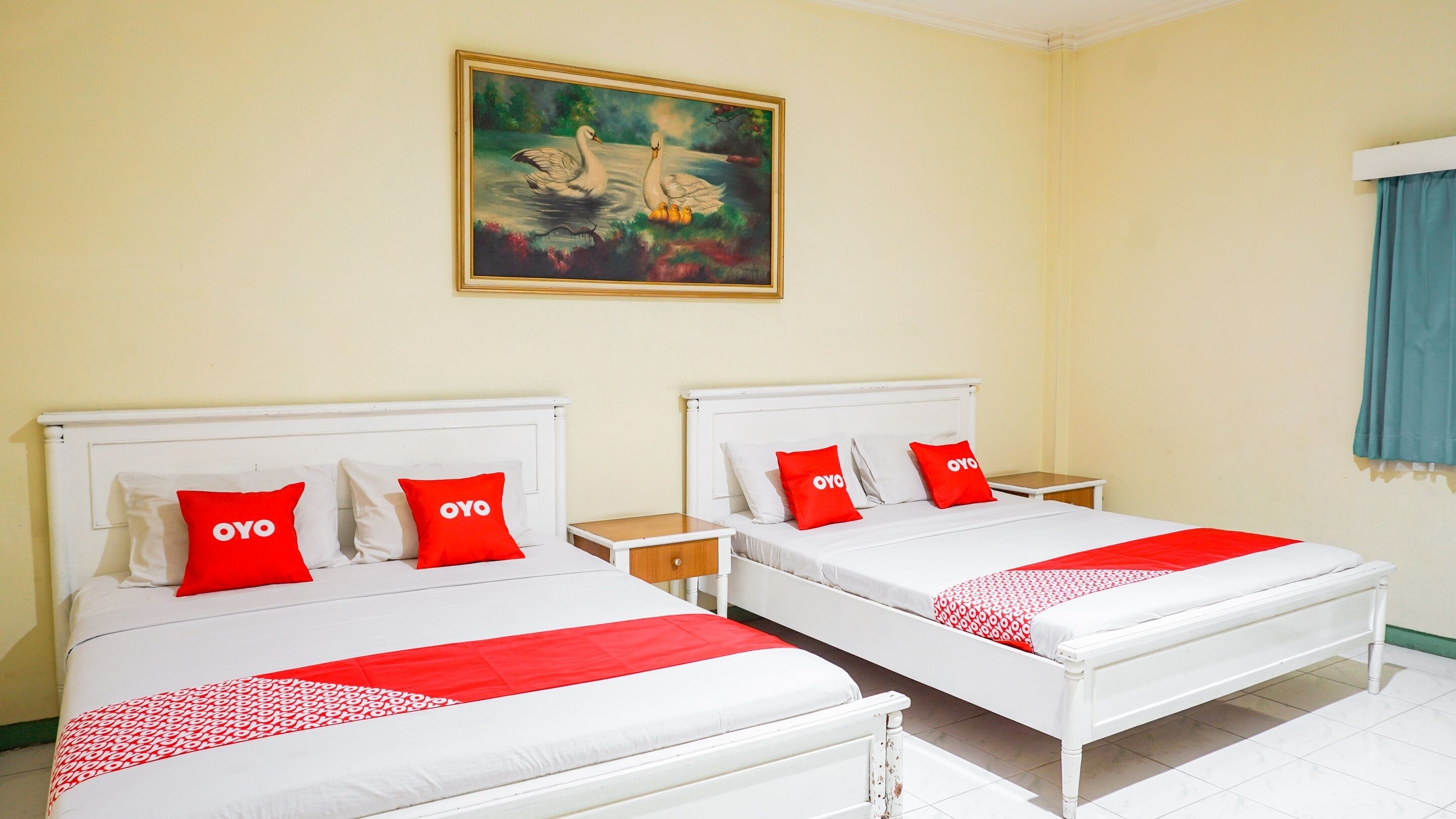 A hotel room with two beds, each with white bedding and red pillows with 'OYO' branding. There is a painting of swans on the wall, and a blue curtain is visible on the right.