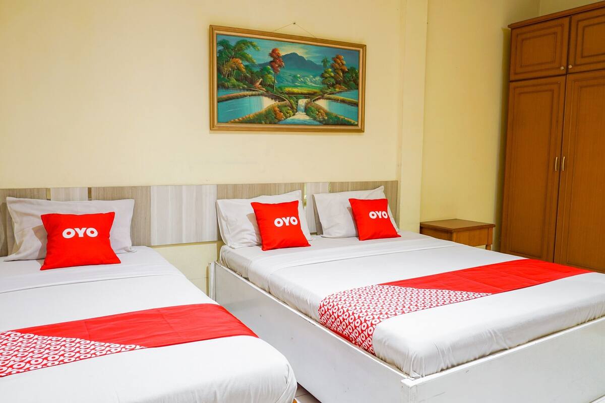 A hotel room with two beds, each with white bedding and red pillows with the word "OYO" on them. There is a painting of a landscape on the wall above the beds, and a wooden wardrobe to the right.